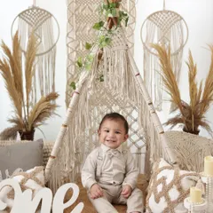 Officially one! A proud and happy one-year-old boy sits in front of a large "one" sign, surrounded by a stylish boho setup with macrame and pampas grass.