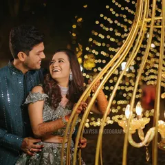 A happy couple shares a laugh during their Sangeet night, with beautiful golden decor and fairy lights creating a magical ambiance.