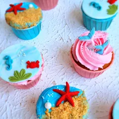 A variety of underwater-themed cupcakes. These show different topping styles, from buttercream swirls with fondant tails to smooth fondant tops with beach scenes.