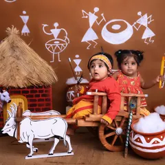 A playful and creative Janmashtami themed shoot. Two babies dressed as little Krishnas sit in a miniature wooden bullock cart, surrounded by a rustic village backdrop, showing the fun and imaginative sets we create.