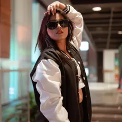 This portrait captures a cool, confident attitude. The model's varsity jacket and sunglasses, combined with the blurred urban background, create a classic street style look.