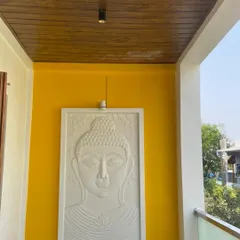 An on-site photo of a completed balcony detail. The vibrant yellow wall provides a cheerful backdrop for a serene white Buddha relief panel, complemented by a modern wooden ceiling, creating a perfect space for reflection.