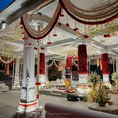Inside the mandap, where pillars are wrapped in dense red roses and the ceiling is draped with garlands of jasmine and red flowers, creating a rich and fragrant canopy.