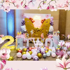 A whimsical butterfly-themed second birthday party featuring large golden butterfly wings as the central photo-op, surrounded by pink, purple, and gold balloons.