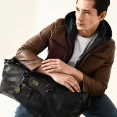 A warm, studio-lit portrait for Kompanero, showcasing their men's leather jacket and duffle bag. The model's relaxed yet confident pose creates an aspirational look for their target customer.