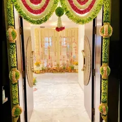 This is a traditional floral entrance for a home event. The doorway is framed with intricate garlands made of fresh flowers and green leaves, looking into the main event space.