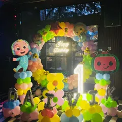 We love creating magical parties for kids, like this vibrant Cocomelon first birthday. The setup includes a colorful balloon arch, character cutouts of JJ and the watermelon TV, a neon sign, and a light-up number '1'.