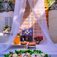 I create picturesque picnic setups perfect for a surprise date. This arrangement includes a classic picnic basket, fresh food and drinks, and a beautiful floral foreground, all framed by a delicate sheer canopy.