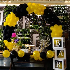 A bold and modern decoration with a black and yellow balloon arch against a green wall. This setup includes a dessert table, floral accents, and transparent 'BABY' blocks filled with matching balloons for a stylish celebration.