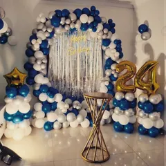 A vibrant 24th birthday decoration featuring a blue, white, and silver balloon arch over a silver foil fringe backdrop, complete with foil number balloons on pillars.
