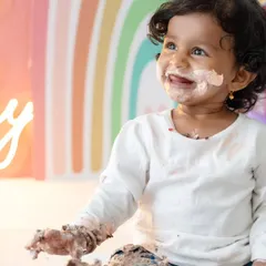 A close-up of a baby girl enjoying her cake smash, with a look of pure delight and a face covered in delicious frosting. The colorful rainbow backdrop adds to the cheerful atmosphere.