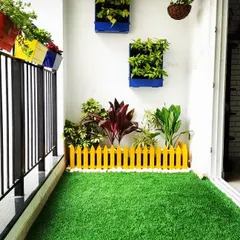 A bright and cheerful balcony floor. The entire space is covered in vibrant artificial grass, with a small yellow picket fence adding a playful touch and separating the potted plants.