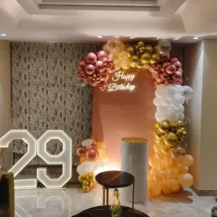 A chic 29th birthday celebration at home. The decor features a modern balloon arch in rose gold, gold, and white against a peach backdrop, with a large light-up number '29'.