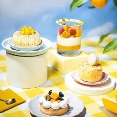 A beautiful assortment of mango desserts, including tarts, mousse, and cheesecakes, styled as a whimsical summer picnic. The tiered arrangement and fresh mango branches create a dreamy, surreal scene.