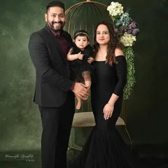 An elegant family of three. The parents and their one-year-old daughter are dressed in black, posing with a golden birdcage prop against a rich green backdrop for a touch of artistic flair.