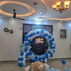 This is a classic "Welcome Boy" decoration featuring a full ring balloon arch in blue, white, and gold. The bright "Welcome Boy" neon sign makes it a perfect photo backdrop for your celebration at home.