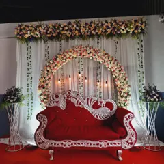 A classic stage decoration for an engagement or reception. The centerpiece is a beautiful floral arch over a royal red and silver sofa, creating a perfect spot for the couple.