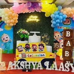 A vibrant and cheerful Cocomelon theme party, perfect for a first birthday. This setup includes character cutouts, a rainbow balloon arch, a custom backdrop, and large marquee letters spelling out the names of the birthday twins.
