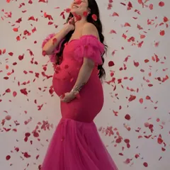 Celebrate your joy with this vibrant hot pink mermaid gown. The off-shoulder ruffle sleeves and form-fitting style beautifully highlight your baby bump, while the falling petals add a touch of playful romance to this studio shot.