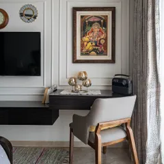 A compact and functional study nook carved out in a bedroom. The floating desk and classic chair are placed beside the TV unit, with traditional Indian art adding a touch of cultural elegance.