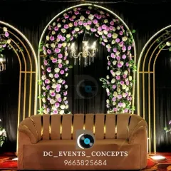 A modern and elegant Mehendi backdrop featuring three golden arches adorned with purple and white flowers. The integrated chandeliers add a touch of glamour to the evening festivities.