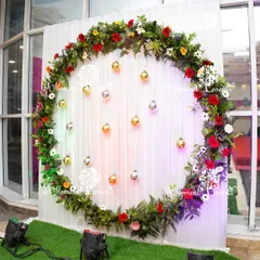 This beautiful photo booth was created for an anniversary celebration, featuring a large circular ring adorned with fresh greenery, red and white flowers. Hanging gold and silver baubles add a touch of sparkle against the clean white curtain backdrop.