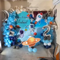An exciting space-themed birthday decoration for a second birthday. This setup includes a custom backdrop, astronaut and rocket cutouts, planet props, and a balloon garland in different shades of blue.