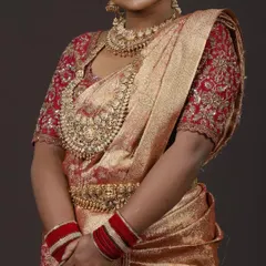 This is a classic South Indian muhurtham look. The rich red and gold Kanchipuram silk saree is paired with a custom-designed blouse featuring intricate zardozi and maggam embroidery, creating a timeless bridal outfit available for rent.