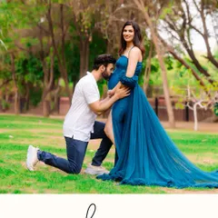 A heartfelt outdoor maternity photo of a partner kneeling to kiss his wife's baby bump. The vibrant blue of her flowing gown stands out against the lush green park, capturing a perfect moment of love and anticipation.