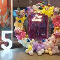 A magical unicorn-themed party for a 5th birthday. This vibrant setup includes a circular balloon arch in pastel rainbow colors, a sparkling pink fringe backdrop, a 'Happy Birthday' neon sign, and a large light-up number '5' for a truly enchanting celebration.