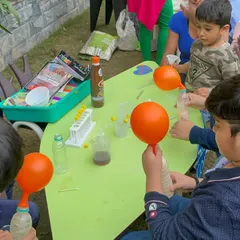 A hands on science experiment in progress. Children are gathered around a table, learning about chemical reactions by inflating balloons with a simple, safe mixture. It's learning by doing.