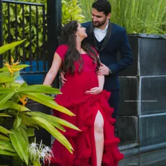 An expecting couple shares a loving gaze in a garden setting. The mother-to-be looks radiant in a ruffled red gown, showcasing a stylish outdoor maternity look.