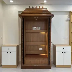 A stunning, full-sized pooja mandir in a dark wood finish. The doors feature traditional jali patterns, and the interior is warmly lit, creating a serene and divine atmosphere. It is flanked by matching storage units.