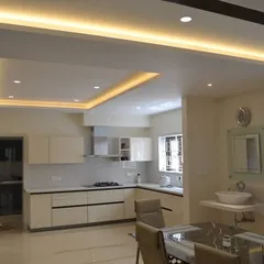 This spacious open-plan area showcases a minimalist all-white modular kitchen that blends seamlessly with the dining space. The design is clean and bright, with layered ceiling lights providing ample illumination for both cooking and dining.