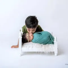 A tender moment as an older brother leans in to give his sleeping newborn sister a kiss. The minimalist white background and simple prop bed keep the focus entirely on this sweet sibling connection.