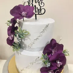 A modern and luxurious two-tier wedding cake. The crisp white fondant is accented with diagonal crystal detailing and cascades of deep purple orchids, complete with a "Mr & Mrs" topper for a sophisticated finish.