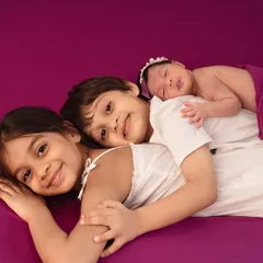 Three siblings snuggled together, with the newest addition resting peacefully. The vibrant purple backdrop adds a pop of color to this sweet and loving newborn and sibling portrait.