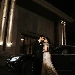 A glamorous outdoor portrait of the couple on their reception night. Posed in front of a luxury car and a grand, well-lit building, this shot has a sophisticated and high-fashion feel.