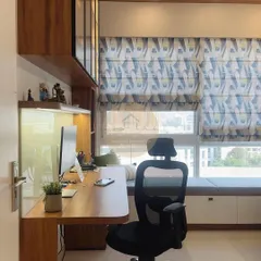 A cozy and functional study cum office space. This design features a long wooden desk, ample storage with overhead cabinets and a window-side bench, and beautiful patterned roman blinds that control natural light.