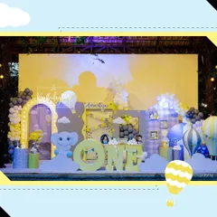 A cheerful and whimsical first birthday celebration with a hot air balloon and baby elephant theme. The complete stage setup features large 'ONE' block letters, custom backdrops, and a soft color palette of yellow, grey, and blue.