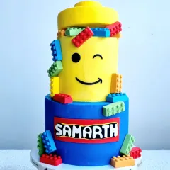A fun, two-tier Lego-themed cake. The cake is designed to look like Lego blocks, with a winking Lego head and colorful fondant bricks.