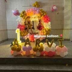A beautiful first birthday setup for Aarvi with a circular balloon arch in pink, white, and gold. The decoration includes a custom photo backdrop, a 'Happy Birthday' neon sign, a light-up number '1', and balloon letters spelling out the name.
