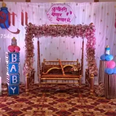 A sweet and simple Dohale Jevan setup featuring a 'कुणीतरी येणार येणार' sign. The decoration includes a floral swing and cute baby pram cutouts for a boy and girl.