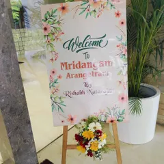 A simple and elegant welcome board for a Mridangam Arangetram. The floral-printed sign is placed on a wooden easel and decorated with a fresh flower arrangement.
