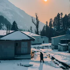 A golden winter sun hangs over a snow-covered village in Aru Valley, Kashmir. The long shadow of a lone walker adds a sense of scale and solitude to this peaceful scene.