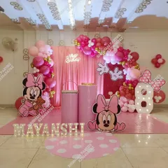 A full-themed Minnie Mouse birthday party decoration for a little one's special day. This setup includes large character cutouts, a pink fabric backdrop, a balloon arch in shades of pink and red, and marquee letters spelling out the birthday girl's name.