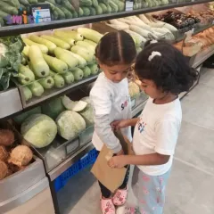 Two children carefully place items in their shopping bag in the fresh produce section. This trip was to procure ingredients for our Independence Day salad, making the learning purposeful.