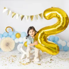 A series of portraits celebrating a second birthday. The toddler poses with a giant gold '2' balloon, surrounded by blue and white balloons and confetti for a festive look.