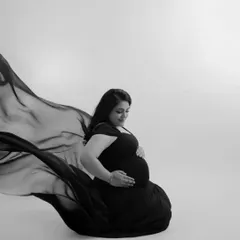 A gentle and introspective black and white portrait. The mother-to-be is seated, looking down at her bump, with the flowing fabric creating a soft, protective circle around her.