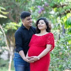 A candid shot of an expecting couple sharing a happy, natural smile. The mother is wearing a beautiful red lace gown, and the sunlight filtering through the trees adds a warm glow.